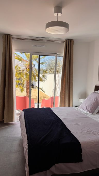 Chambre vue piscine Chambre lumineuse avec lit, rideaux clairs et balcon sur jardin tropical.