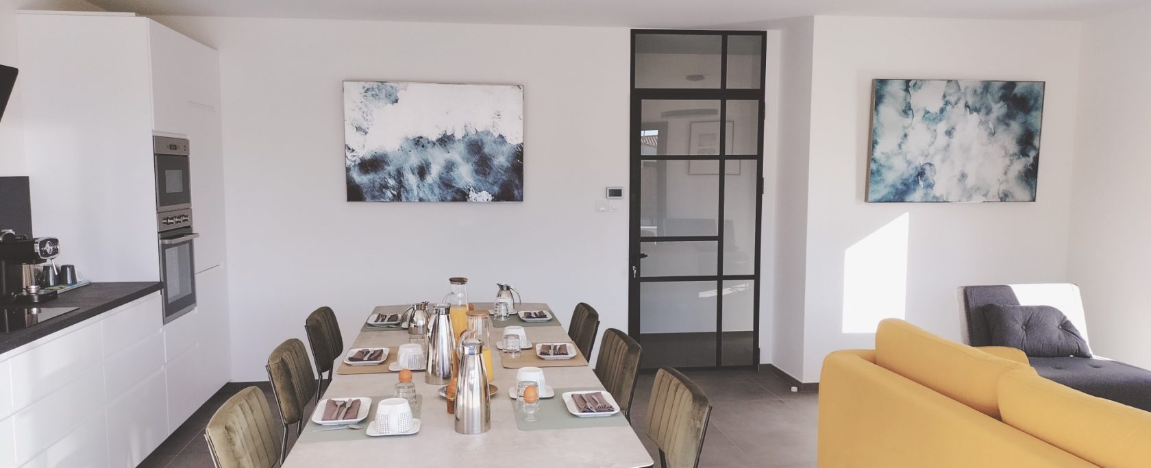 Salle à manger moderne avec une table dressée, canapé jaune et œuvres d'art aux murs.